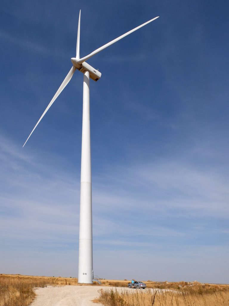 Electric wind turbine with a car at its foot contrasting its enormous size, on a blue sky background