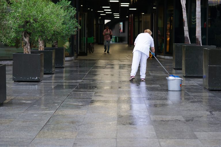 A person wearing white clothing is mopping a shiny, wet floor in a corridor lined with large potted plants. Another person is walking towards the camera in the background under the lit passageway.