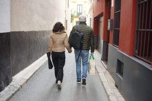 A couple walking hand in hand down a narrow street. The person on the left is wearing a tan jacket and dark pants, holding a rolled item, while the person on the right is wearing a green jacket and jeans, carrying a bag with green handles.