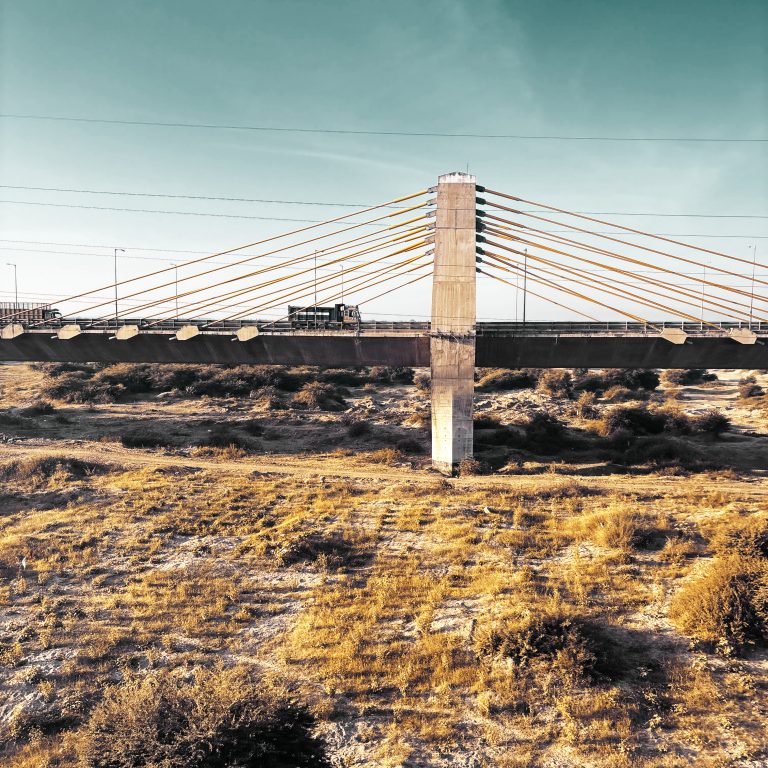 A photo of a cable-stayed bridge spanning a dry, arid landscape. The bridge features a main pillar with numerous cables extending to support the structure