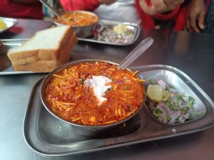 A plate of spicy, noodle-based soup (Kolhapuri Misal) garnished with a dollop of yogurt and chopped onions, served on a metal tray with a side of sliced lemon, cilantro, and additional chopped onions. There are a few slices of bread beside it, and people in the background.