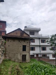 A contrast of architectural styles is depicted in the image: on the left, an old, weathered house made of bricks and mud with a wooden door shows signs of aging and decay, while on the right, a modern white multi-story building with several windows stands in stark contrast. The foreground features a patch of green vegetation amidst a grassy area