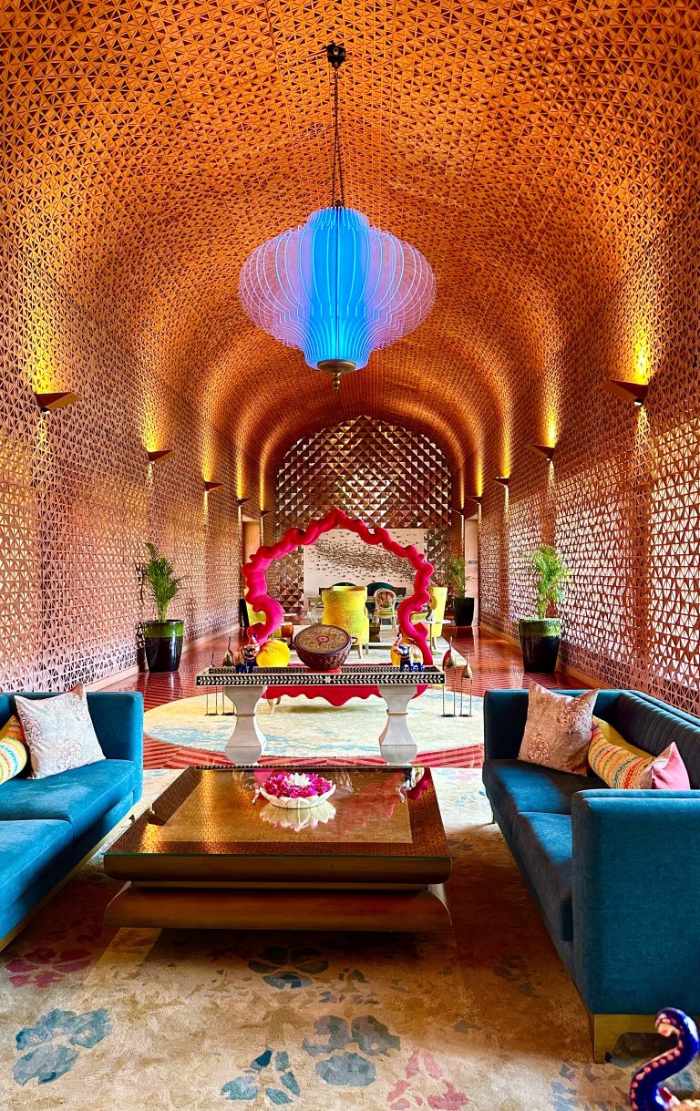 An intricately designed room with geometric patterns on the walls and ceiling, featuring a large blue pendant light. The space includes plush blue sofas with colorful pillows, a central ornate table with decorative items, vibrant accents, and potted plants.