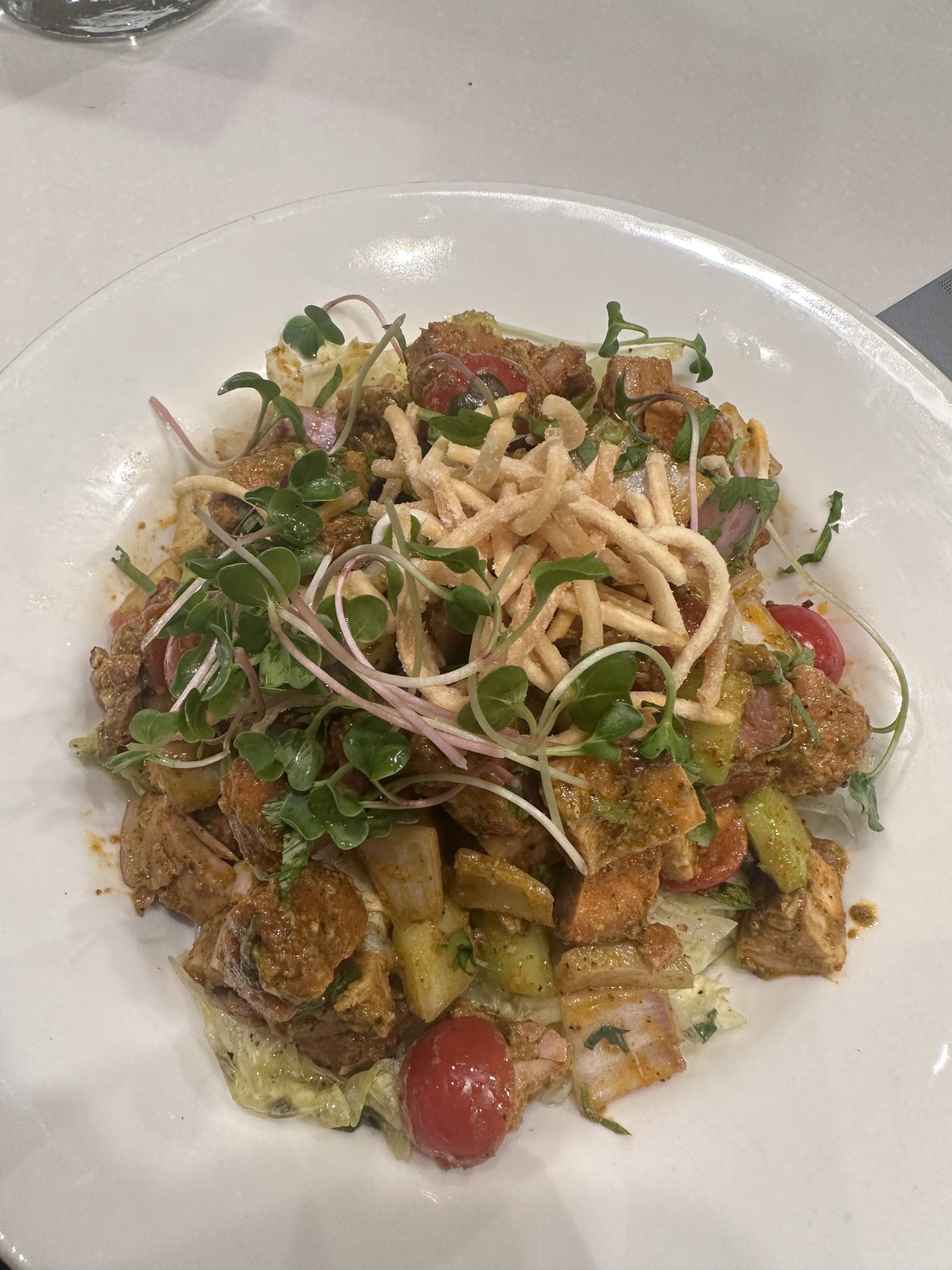 A plate of mixed salad featuring diced chicken, cherry tomatoes, cucumber, microgreens, and crispy fried noodles on top, all lightly dressed and arranged on a white plate.