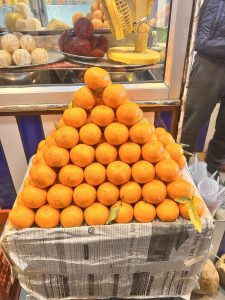 A pyramid stack of fresh oranges displayed on a newspaper-covered box. In the background, there are peeled and whole fruits, along with a machine, possibly a fruit juicer. A person is partially visible wearing a dark-colored outfit.