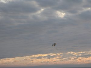 A person is parasailing under a multicolored parachute against a backdrop of a cloudy sky with a faint orange glow near the horizon