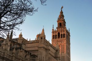 A detailed view of La giralda, a historic tower with intricate architectural features and a weather vane on top, set against a clear blue sky, with bare tree branches framing the scene.