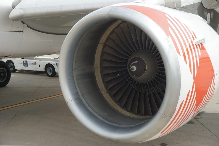 Close-up of a commercial aeroplane jet engine on the tarmac, with visible fan blades and orange striped design on the engine casing. A ground support vehicle is partially visible in the background.