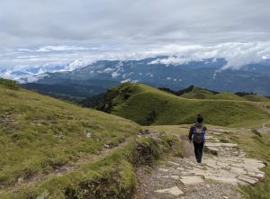 A boy walking in a stairs road of a hilly region. There is a view of the mountains infront of it. The part of mountains is covered with clouds. 