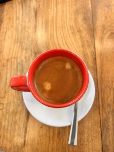 A red coffee cup filled with espresso sits on a white saucer with a metal spoon on a wooden table.