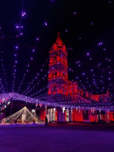 A church tower illuminated in red, surrounded by festive strings of purple lights and decorative stars, with a well-lit canopy structure in the foreground. The scene is set against a dark night sky.