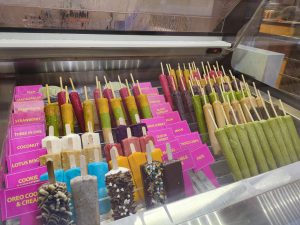 A display case filled with a variety of colorful ice cream sticks, each with different flavors indicated by labels. The ice creams are arranged in neat rows and include options like coconut, strawberry, oreo cookie, and guava sorbet, among others. Some sticks are decorated with chocolate and sprinkles.