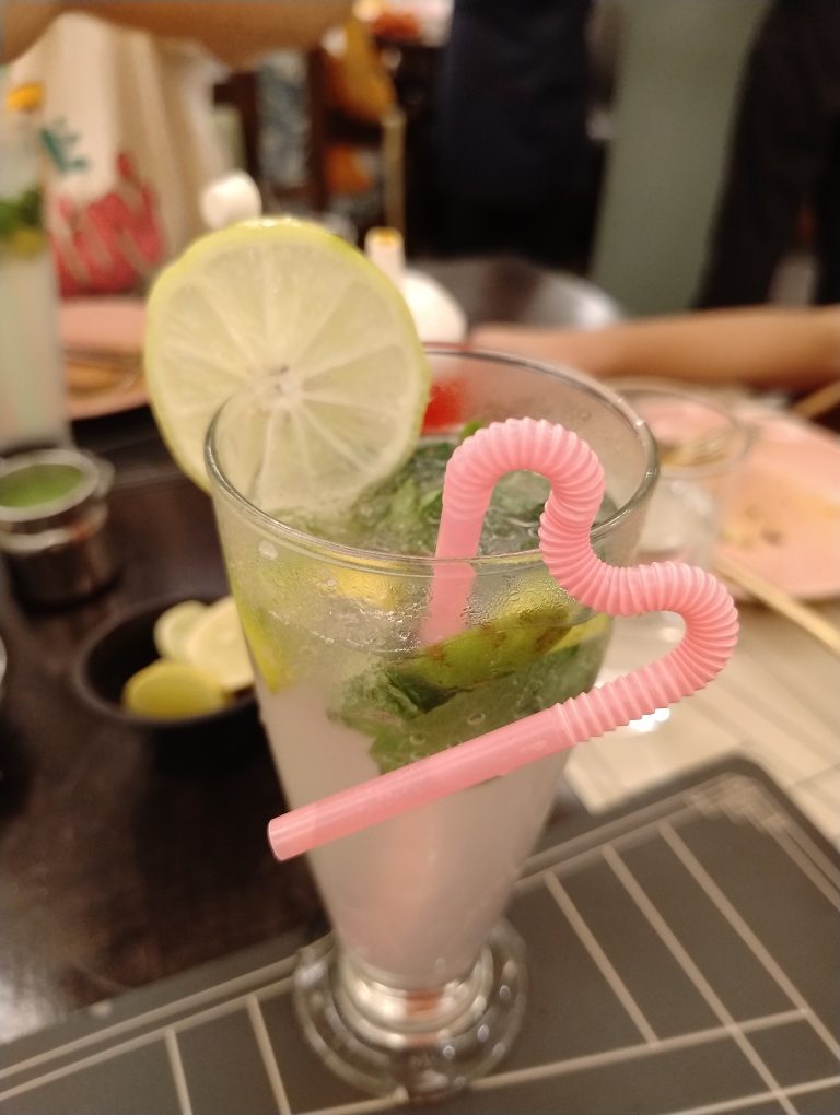 A refreshing beverage in a tall glass, garnished with a slice of lime and mint leaves. The drink has a light, fizzy appearance and is served with a pink, heart-shaped bendy straw. The background shows a dining setting with other plates and glasses blurred in the distance.