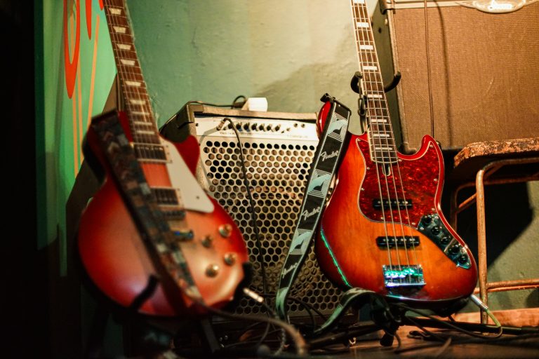 Two electric guitars resting against a wall, with an amplifier in the background. The guitars have colorful, glossy finishes, and the amplifier has a grid-patterned front panel.