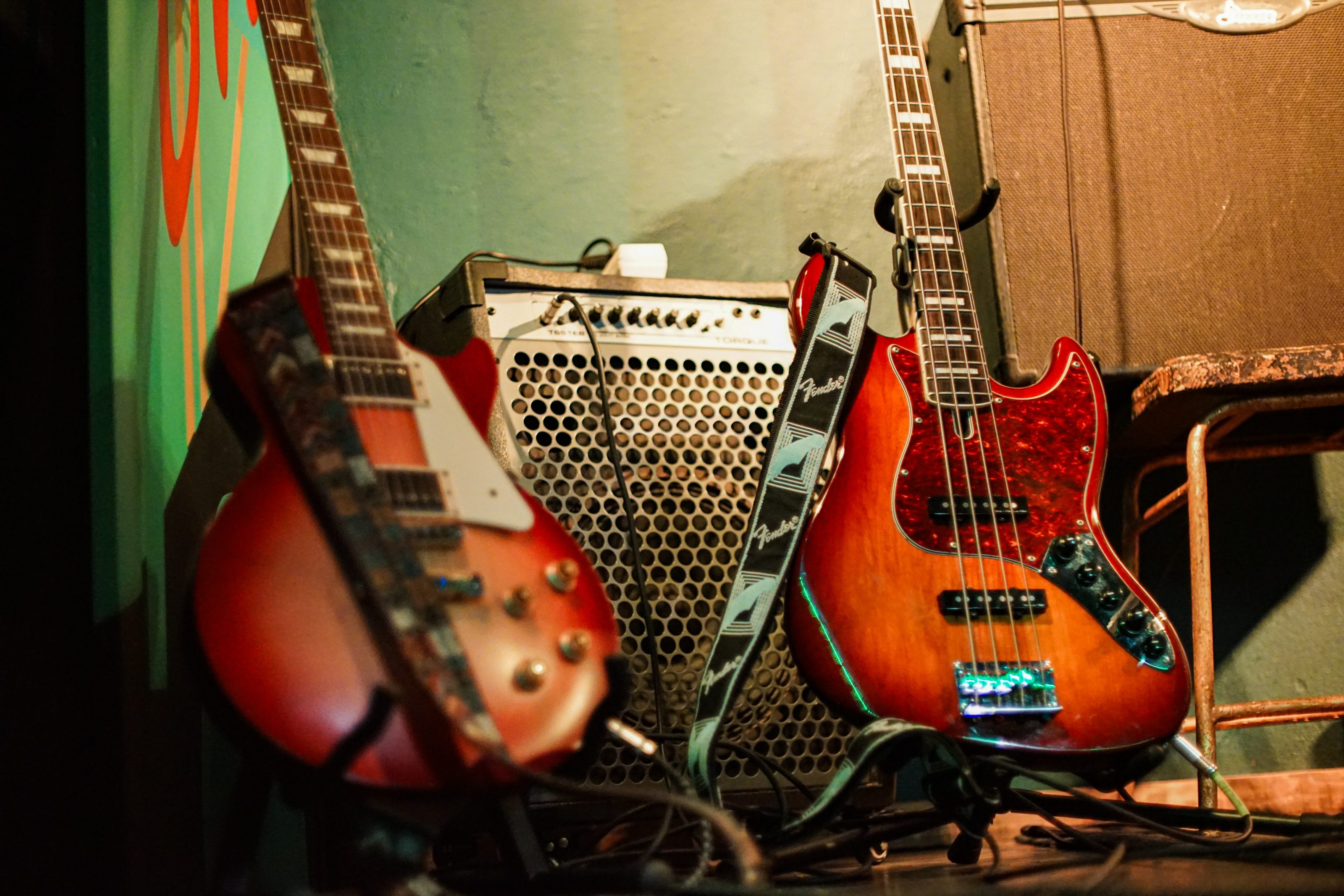 Two electric guitars resting against a wall, with an amplifier in the background. The guitars have colorful, glossy finishes, and the amplifier has a grid-patterned front panel. 