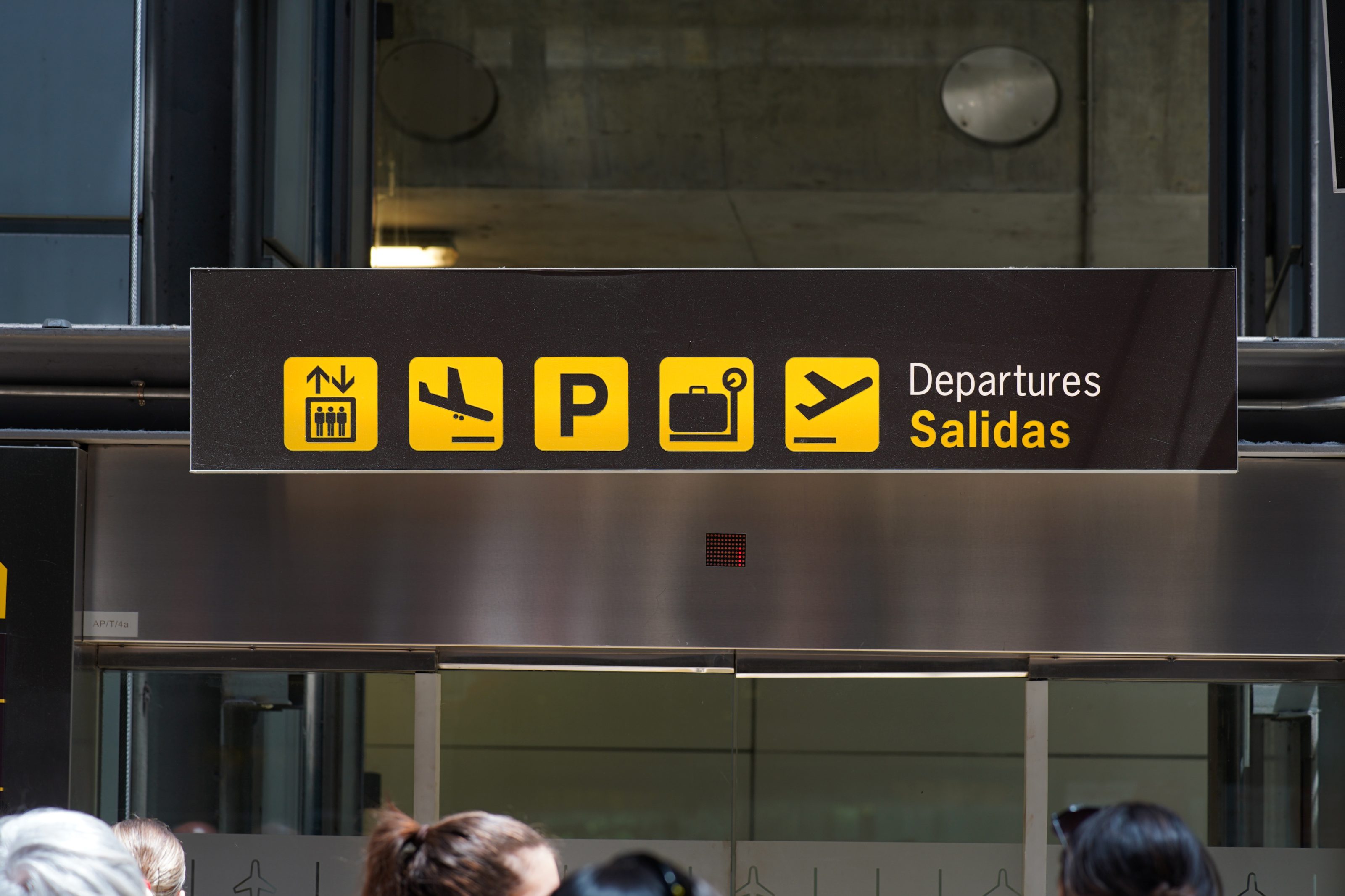 A brown signboard at an airport with yellow symbols indicating directions for elevators, flights, parking, luggage, and departures. The text reads "Departures Salidas." A few people's heads are visible at the bottom of the image.
