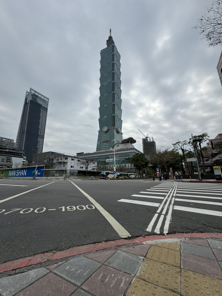A large intersection in Taipei, Taiwan. The Taipei 101 building and a group of people waiting to cross the street are visible in the background.
