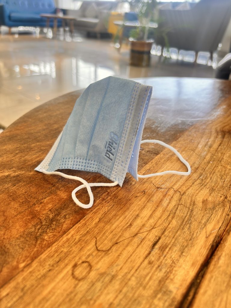 A blue disposable face mask resting on a wooden table in a bright room, with blurred chairs and a plant in the background.