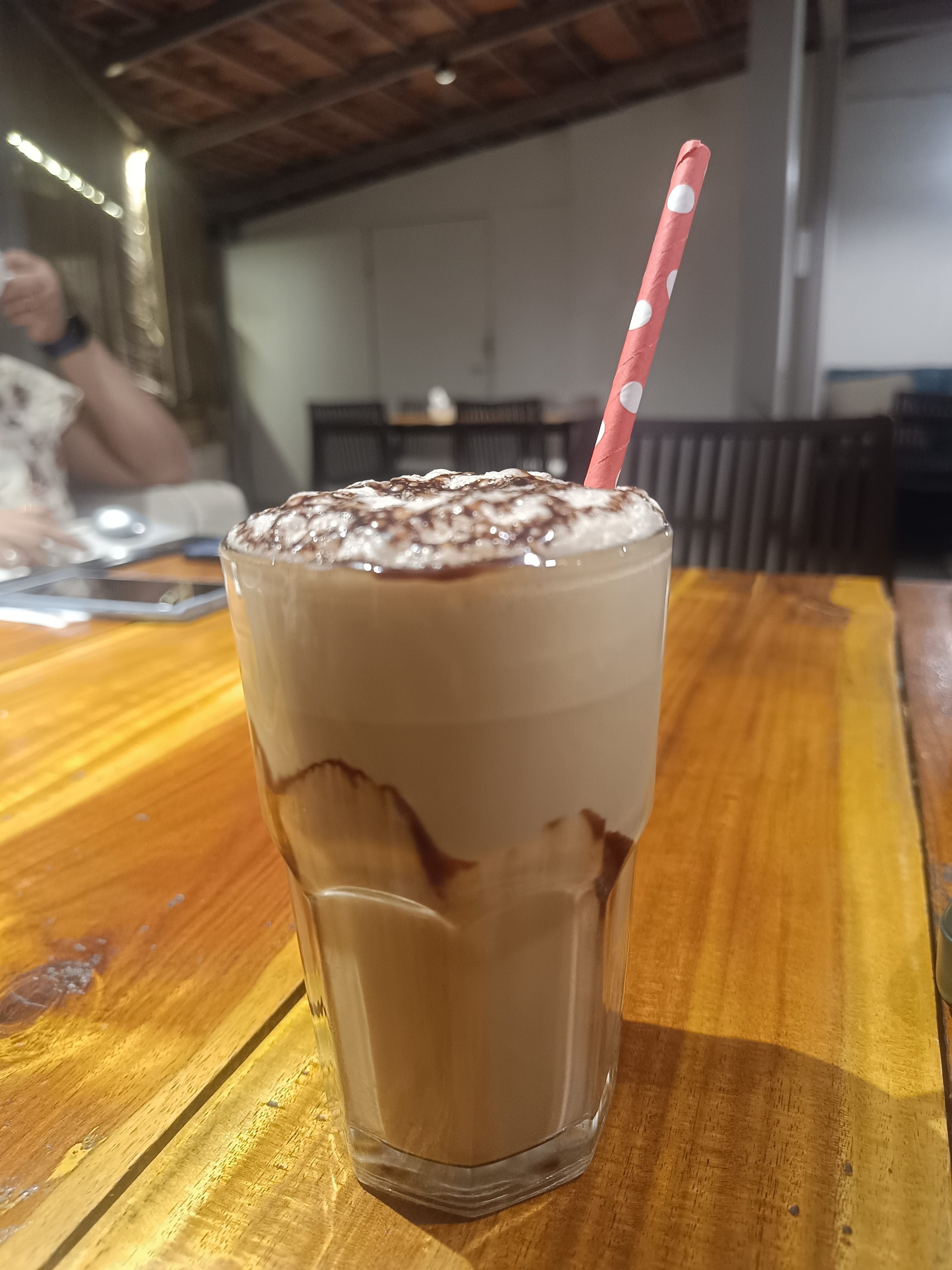 A glass of iced coffee with whipped cream on top, drizzled with chocolate syrup, and a red and white polka dot straw. The drink is on a wooden table, with a blurred background featuring a black bottle and another person partially visible.