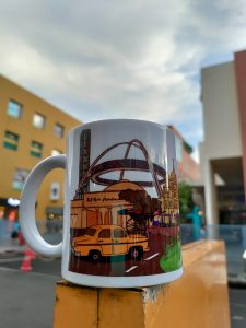 A white mug with colorful illustrations depicting urban scenes placed on a yellow surface, outdoors. Buildings and cloudy sky are visible in the blurred background.