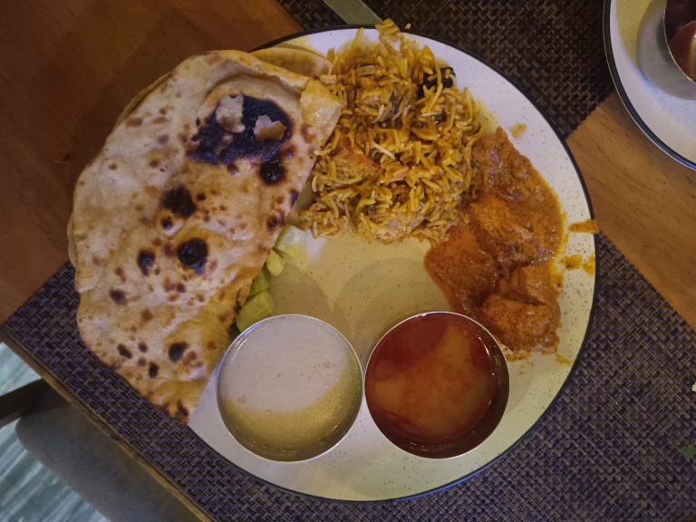 A plate of Indian food featuring naan bread, biryani rice, curry, and two small bowls of sauces on the side. The food is arranged on a white plate placed on a woven placemat on a wooden table.