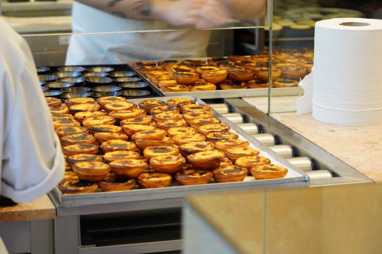 A pastry shop kitchen displaying trays of freshly baked pastéis de nata (custard tarts). A roll of paper towels is visible on the right side of the counter.