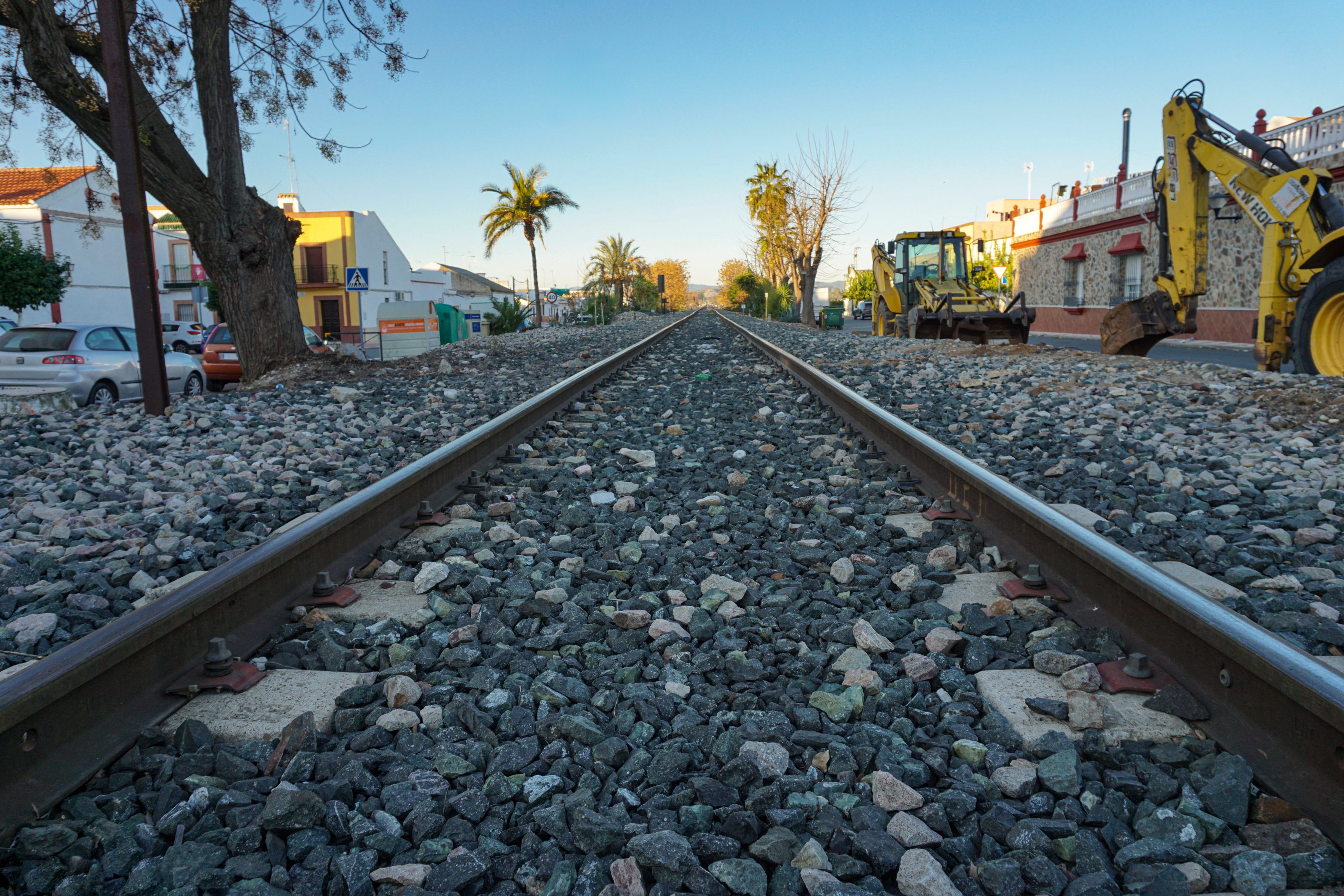 A straight railway track extends into the distance, surrounded by gravel. On the left side, there are parked cars and houses, while on the right side, there are construction vehicles and additional buildings. 