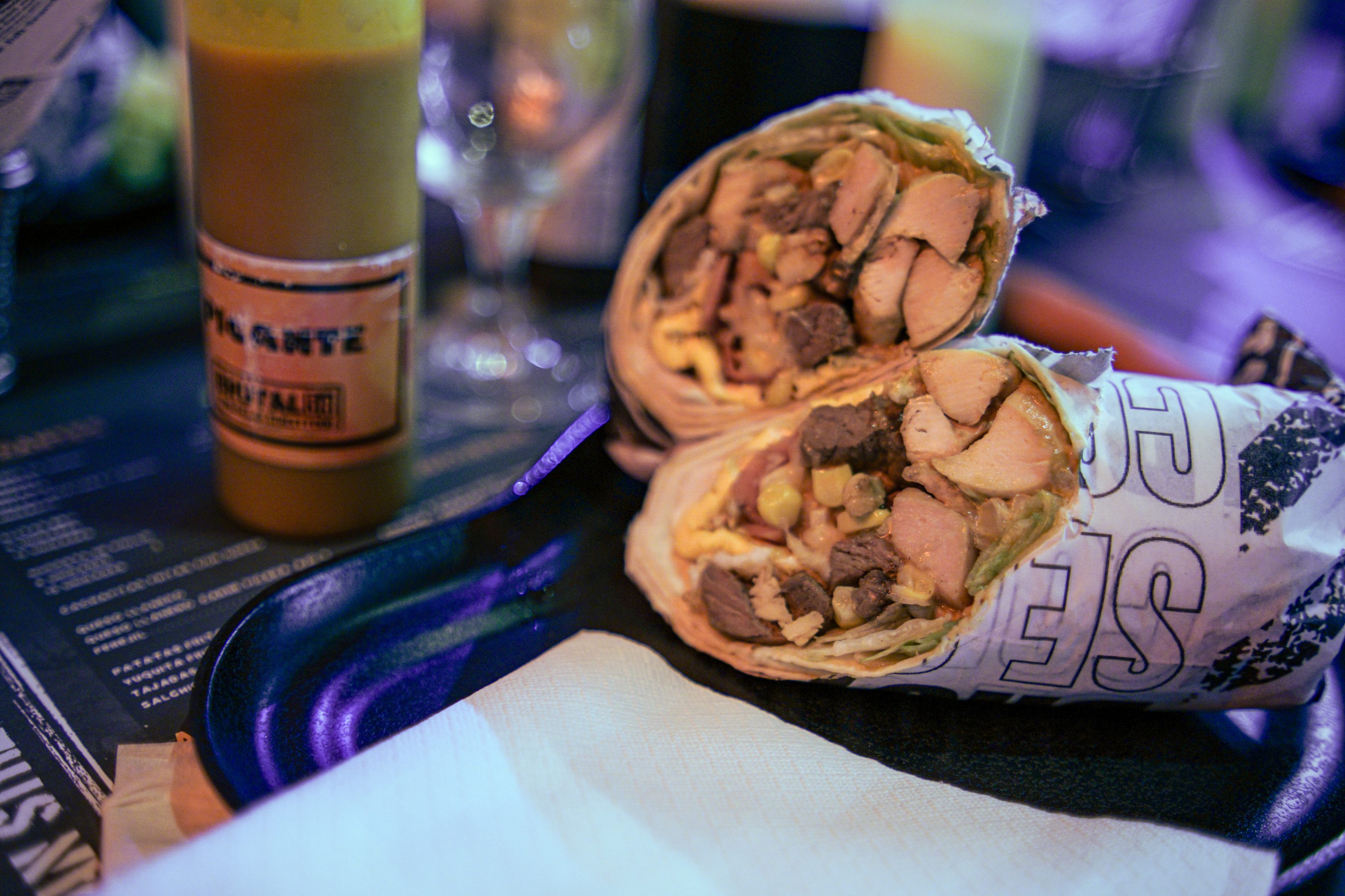 A close-up of a sliced wrap filled with grilled chicken, beef, corn, lettuce, and other ingredients, placed on a tray with a napkin. A bottle of sauce is in the background.