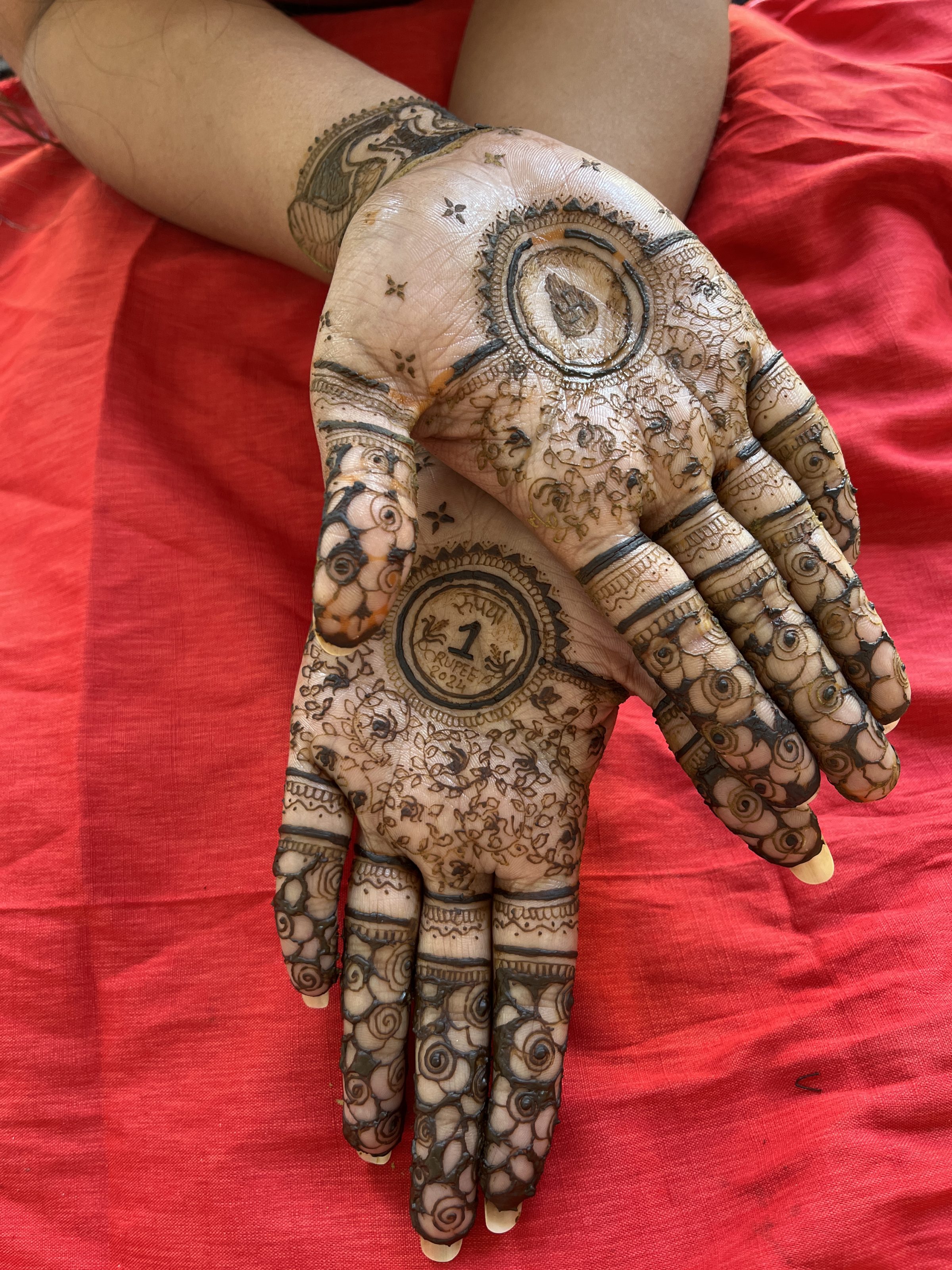 Hands adorned with intricate henna mehndi designs featuring floral patterns and elaborate circular motifs. The background is a red fabric.