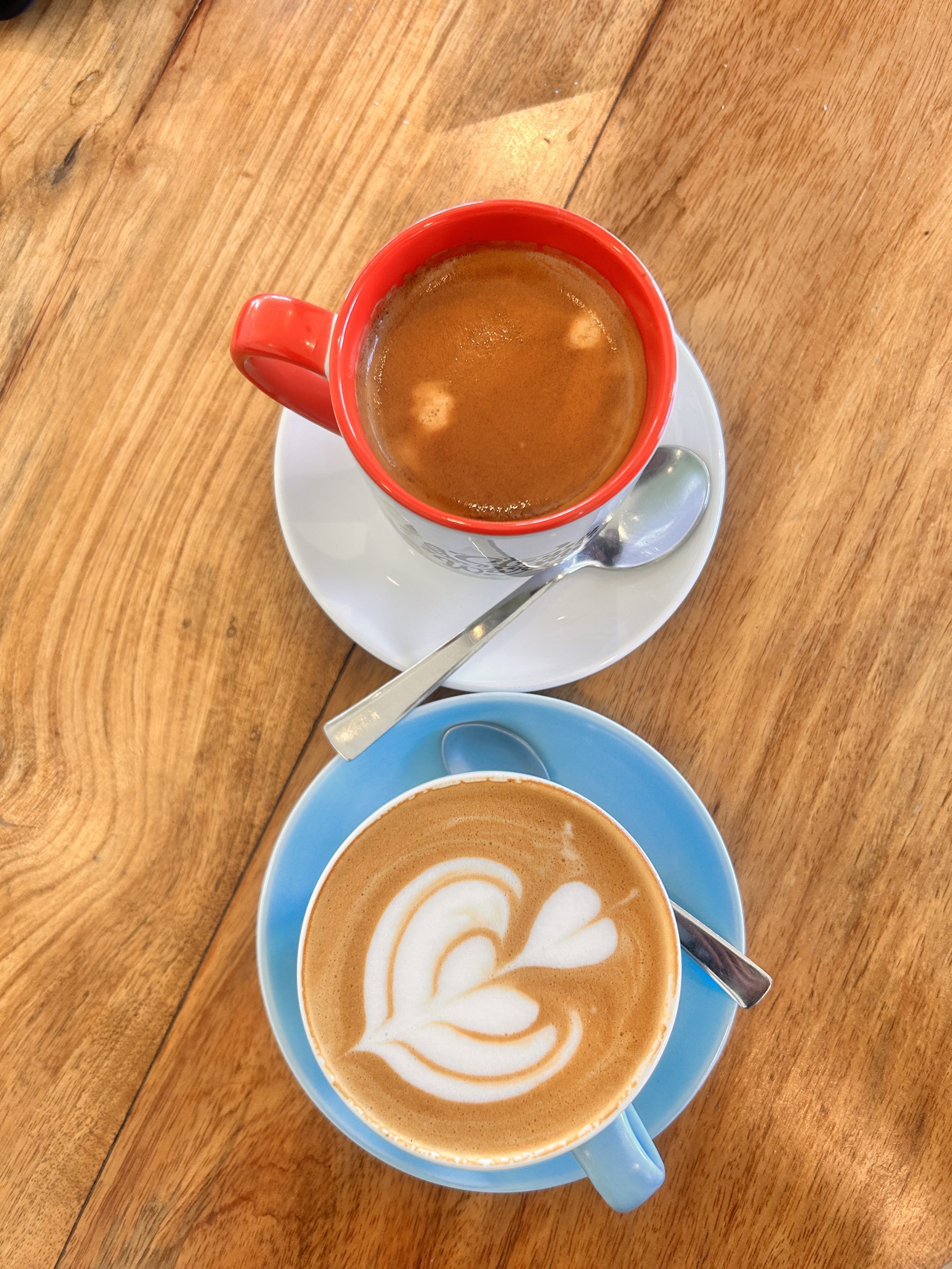 Two cups of coffee on a wooden table, one in a red mug with no visible design and the other in a blue cup with latte art. 