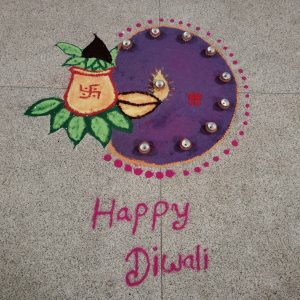 A colorful rangoli design on a stone floor featuring a purple circular pattern with small clay lamps (diyas) arranged around it. On the left, there is a yellow pot with green leaves and a red swastika symbol. Below the design, the words "Happy Diwali" are written in pink.