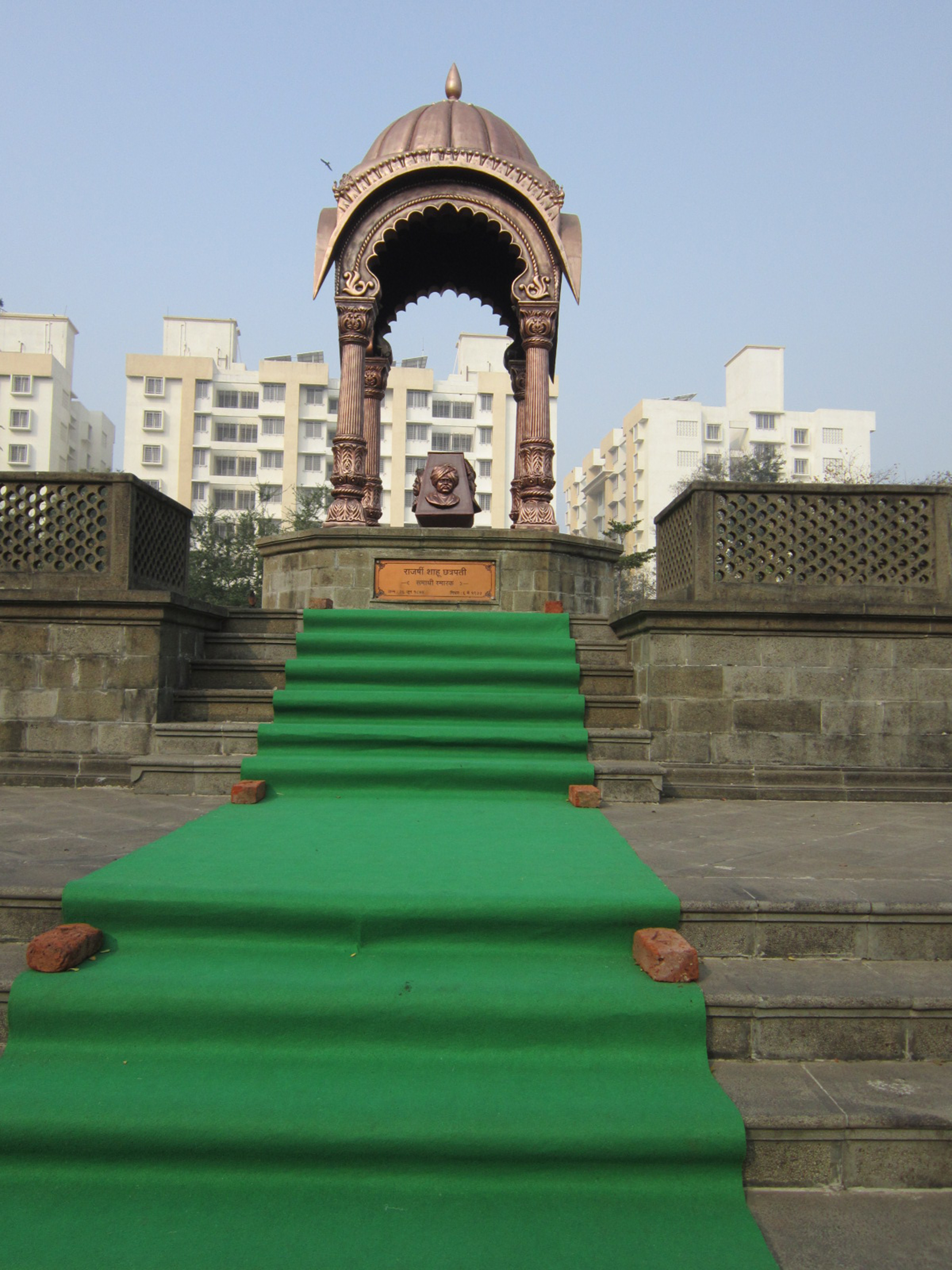 The Rajshri Shahu Maharaj Samadhi in Kolhapur is marked by a bust under a canopy, with a green-carpeted stairway leading up to it and modern buildings in the background.