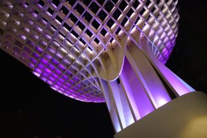 Underside view of a modern architectural structure (Metropol Parasol) illuminated with purple and white lights against a dark sky. The design features a curved, lattice pattern creating an eye-catching geometric effect.