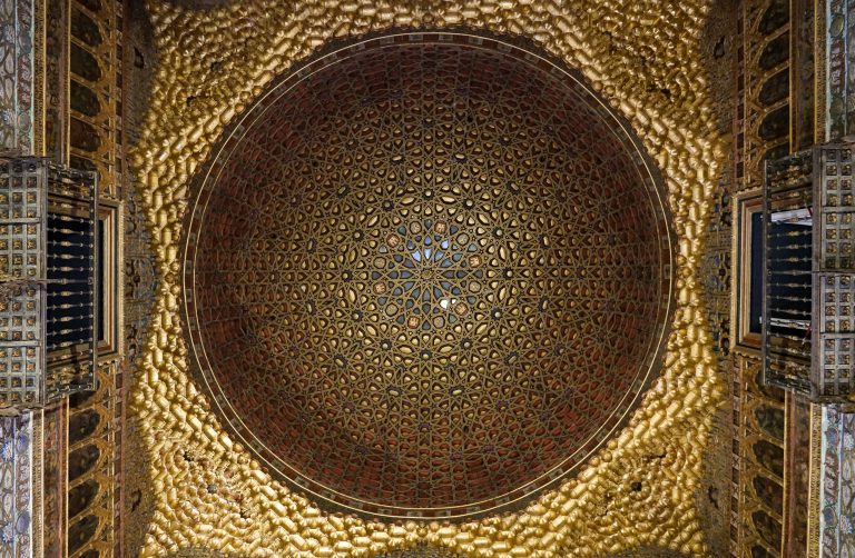 Intricate geometric and floral patterns adorn an ornate domed ceiling, with gold and red hues adding depth and richness. The design is highly detailed and symmetrical, creating a visually stunning focal point.