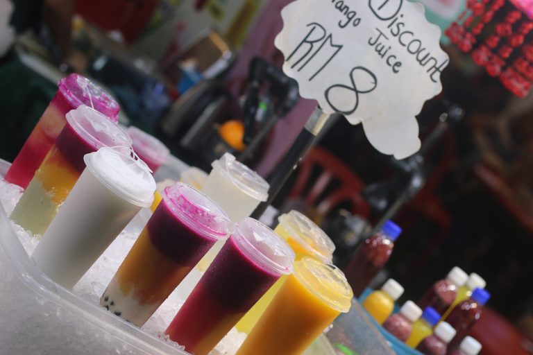 Colorful layered fruit juices in clear plastic cups with lids, displayed on ice, alongside a handwritten sign reading ‘Discount Mango Juice RM 8, A variety of vibrant fruit drinks, including dragon fruit and mango flavors, in cups placed on a bed of ice at a market stall.