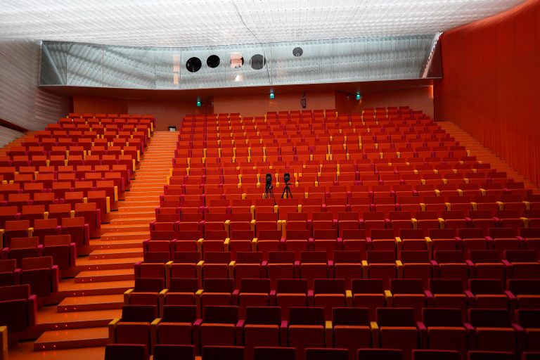 An empty auditorium with several rows of red and orange seats. Two cameras on tripods are set up in the middle of the seating area.