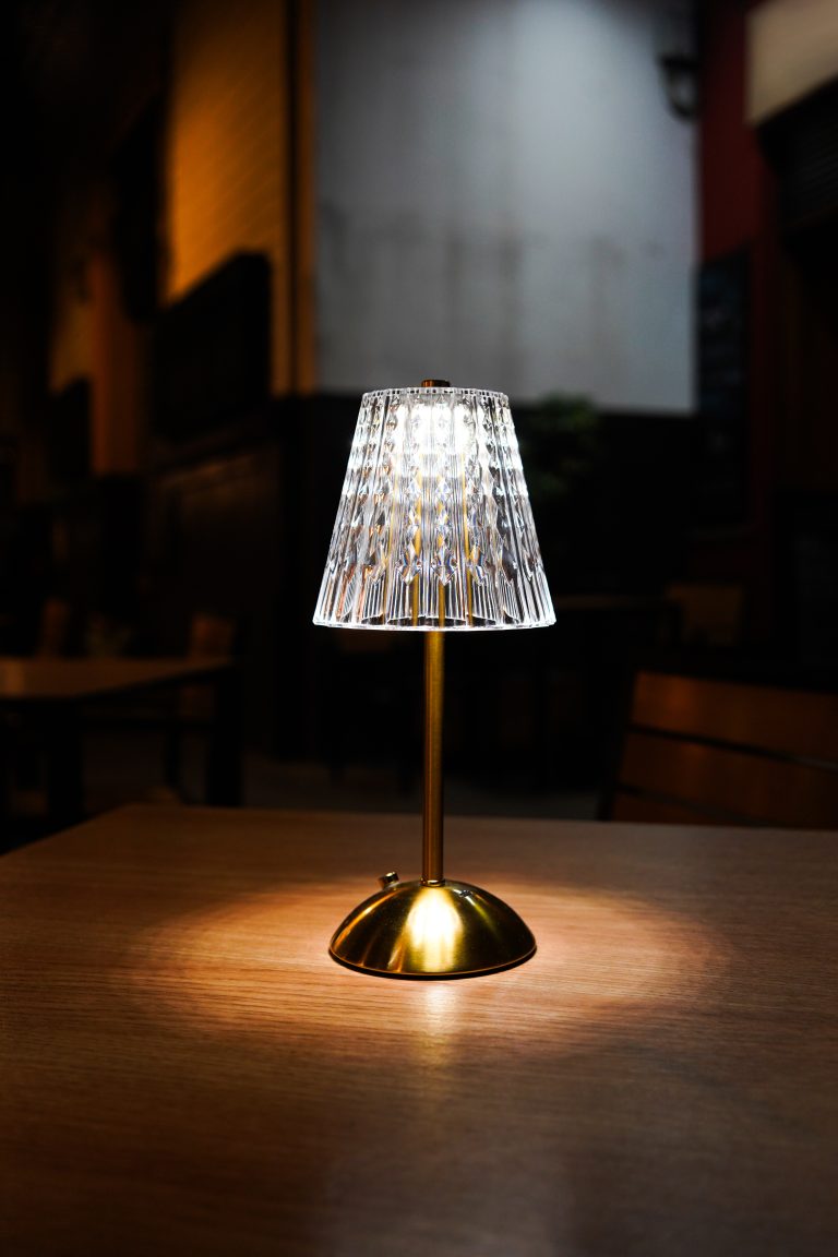 A table lamp with a patterned glass shade and a golden base is illuminated on a wooden restaurant table in a dimly lit street.