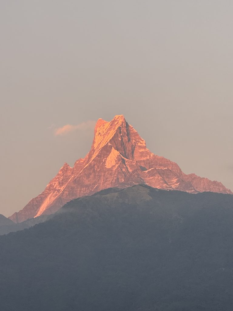 A majestic Mt Fishtail peak bathed in warm, golden sunlight under a clear sky. The foreground is a darker silhouette of rolling hills.