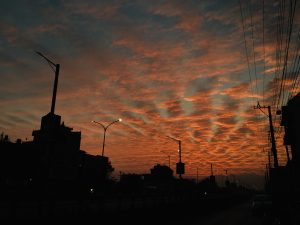 A vibrant sunset sky with orange and red hues over a silhouette of streetlights and buildings. Power lines are visible against the colorful sky, creating a dramatic urban scene.