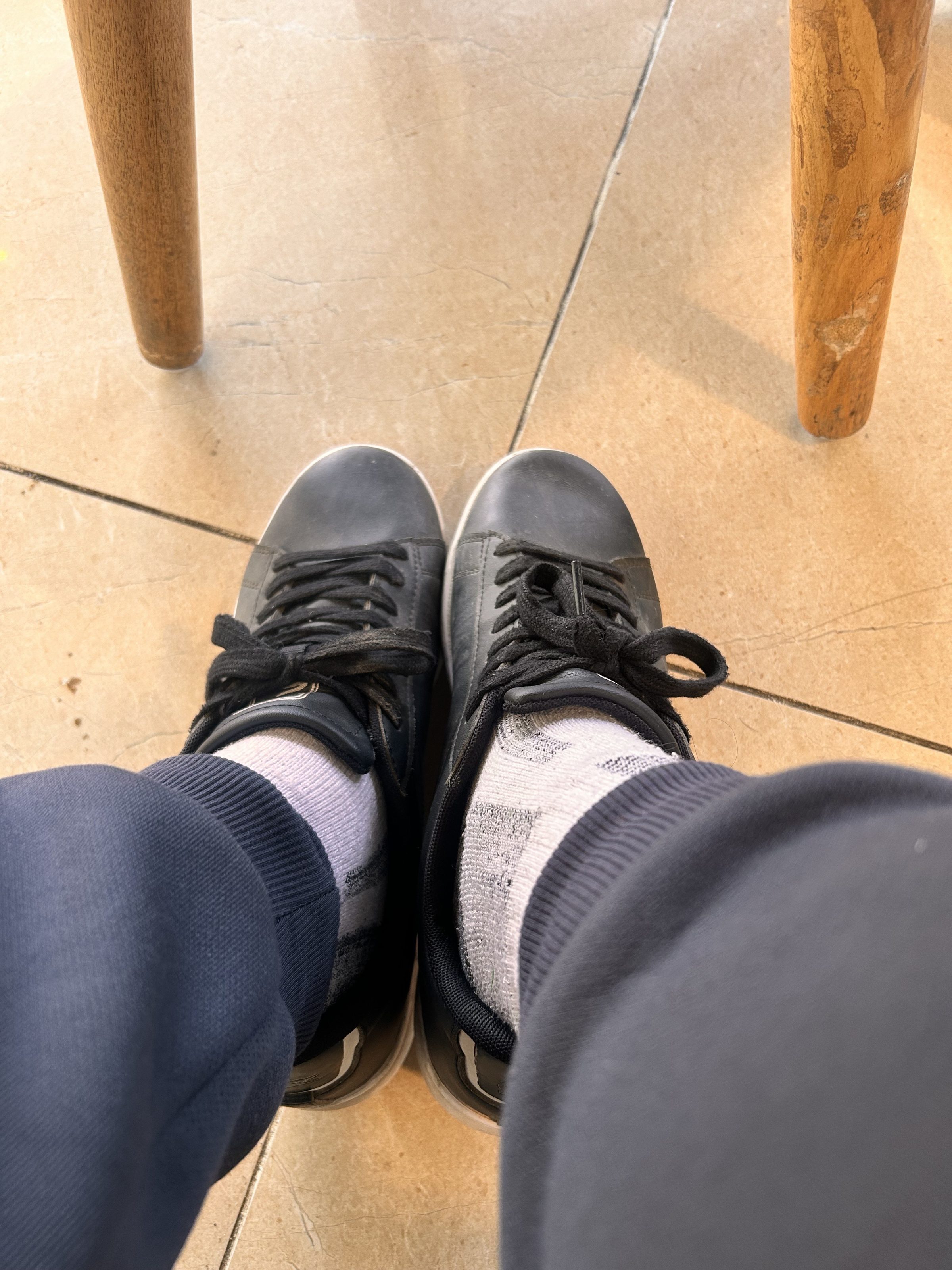 A person wearing black sneakers with white patterned socks and dark pants, sitting on a tiled floor next to wooden table legs.