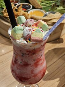 A glass full of ice-cream and strawberry on the top and some chocolate. A bucket full of starter items in the back. 