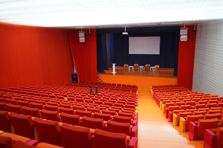 An empty auditorium with rows of orange and red seats facing a stage. The stage has four chairs, a small table, and a podium, with a large projection screen above. The walls are covered with red curtains, and two cameras are set up in the seating area.