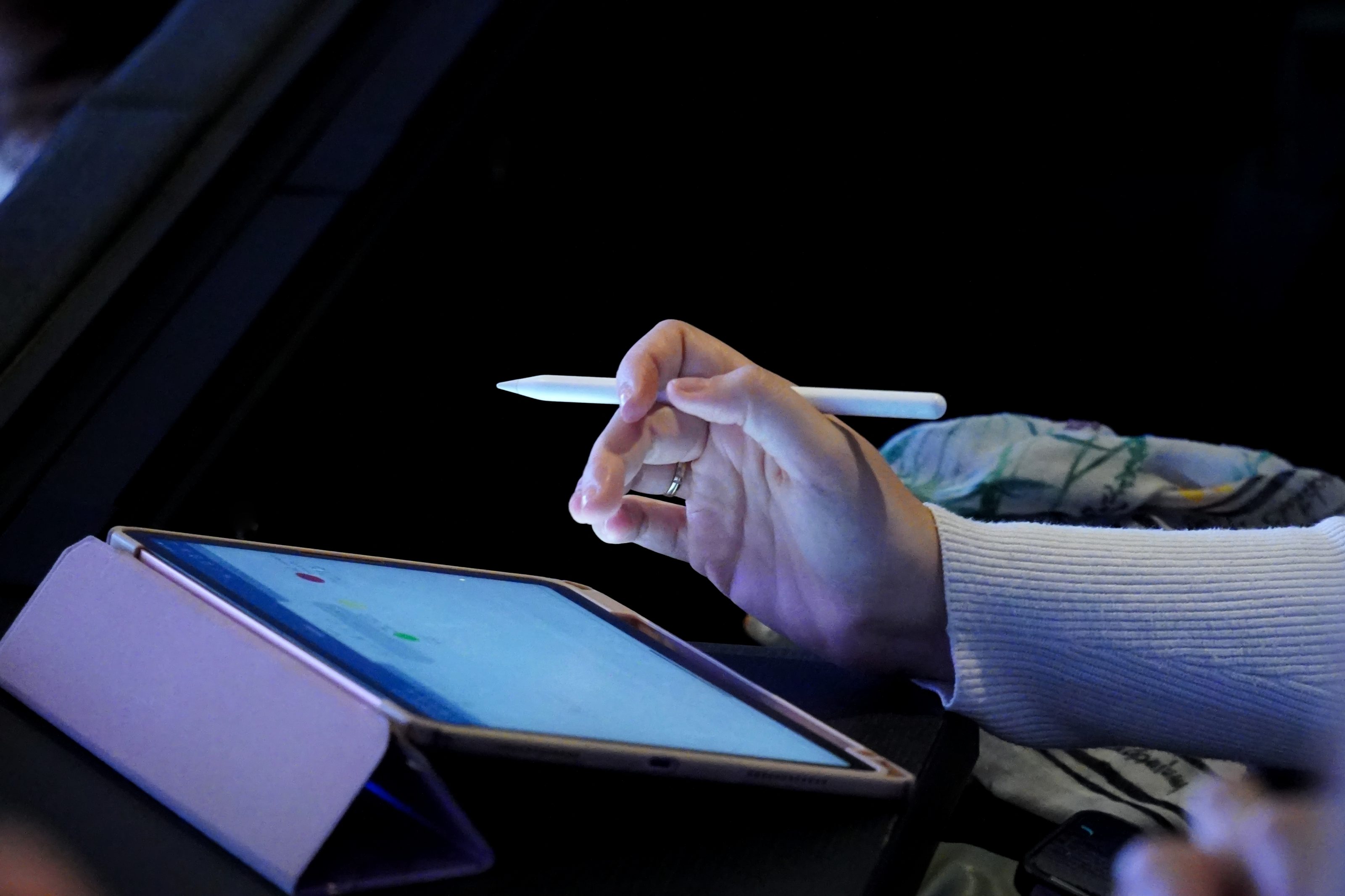 A person holding a stylus over a tablet screen, with the tablet resting on a case. The person is wearing a light-colored, ribbed long-sleeve shirt, and the background is dark.
