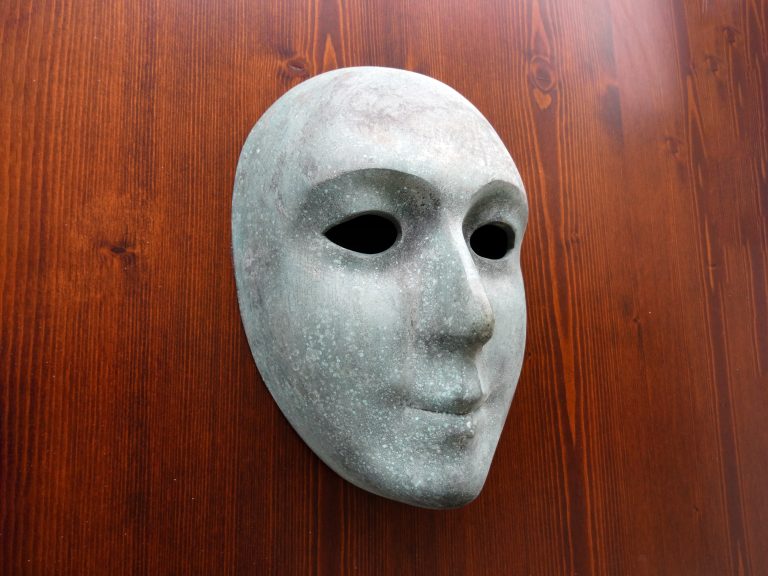 Mask with a smooth, expressionless face is mounted on a wooden background. The mask has hollow eyes and a subtle stone texture.