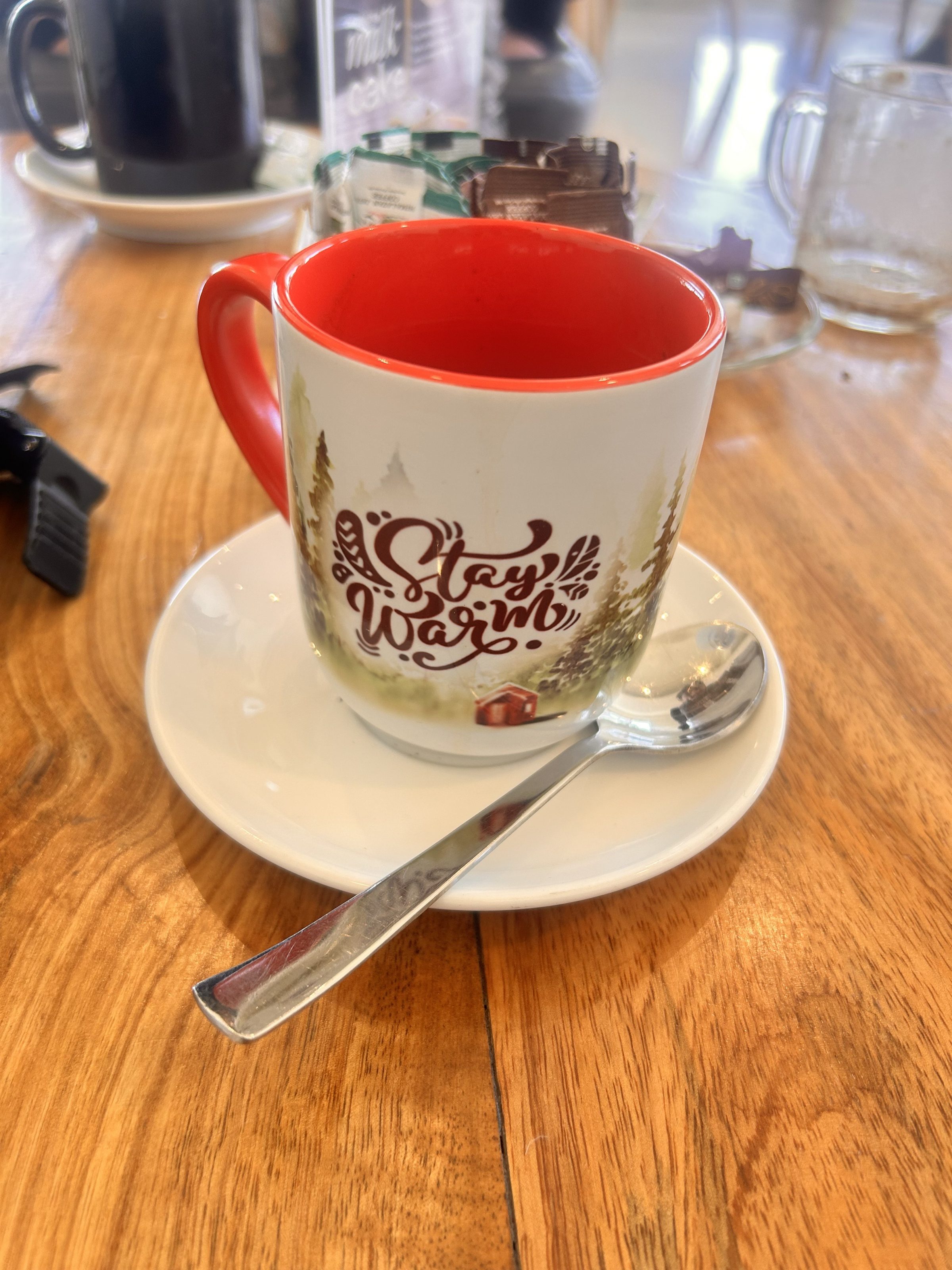 A white mug with a red interior and handle, featuring a design with trees and the words "Stay Warm." 