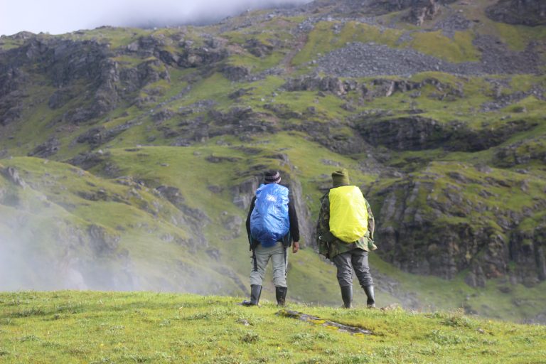 Two hikers wearing backpacks, one covered with a blue rain cover and the other with a yellow cover, stand on a grassy hillside looking towards a mountainous landscape with rocky terrain and patches of green vegetation.