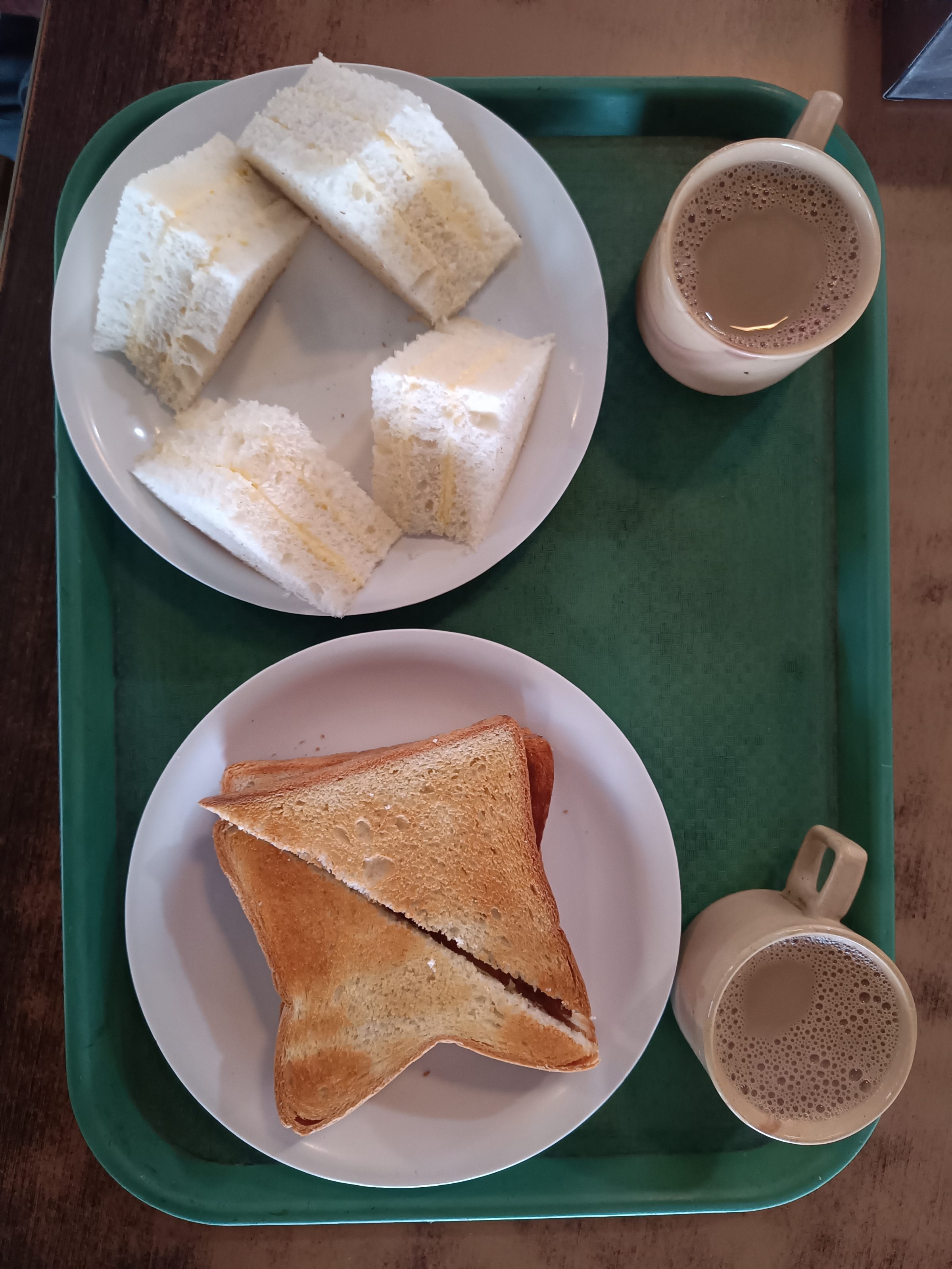 A green tray holds two plates of sandwiches and two mugs of coffee. The top plate contains four triangular sandwiches with white bread and a butter spread. The bottom plate has two slices of toasted bread cut diagonally. Each plate is accompanied by a mug of coffee.