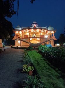 A beautifully illuminated Vivekananda asharam – belagavi at night with a prominent Om symbol on top, surrounded by a neatly maintained garden with grass and potted plants along a paved pathway.
