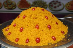 A mound of vibrant orange-hued sweetened grains, garnished with red, glossy dried fruits, on a silver platter.
