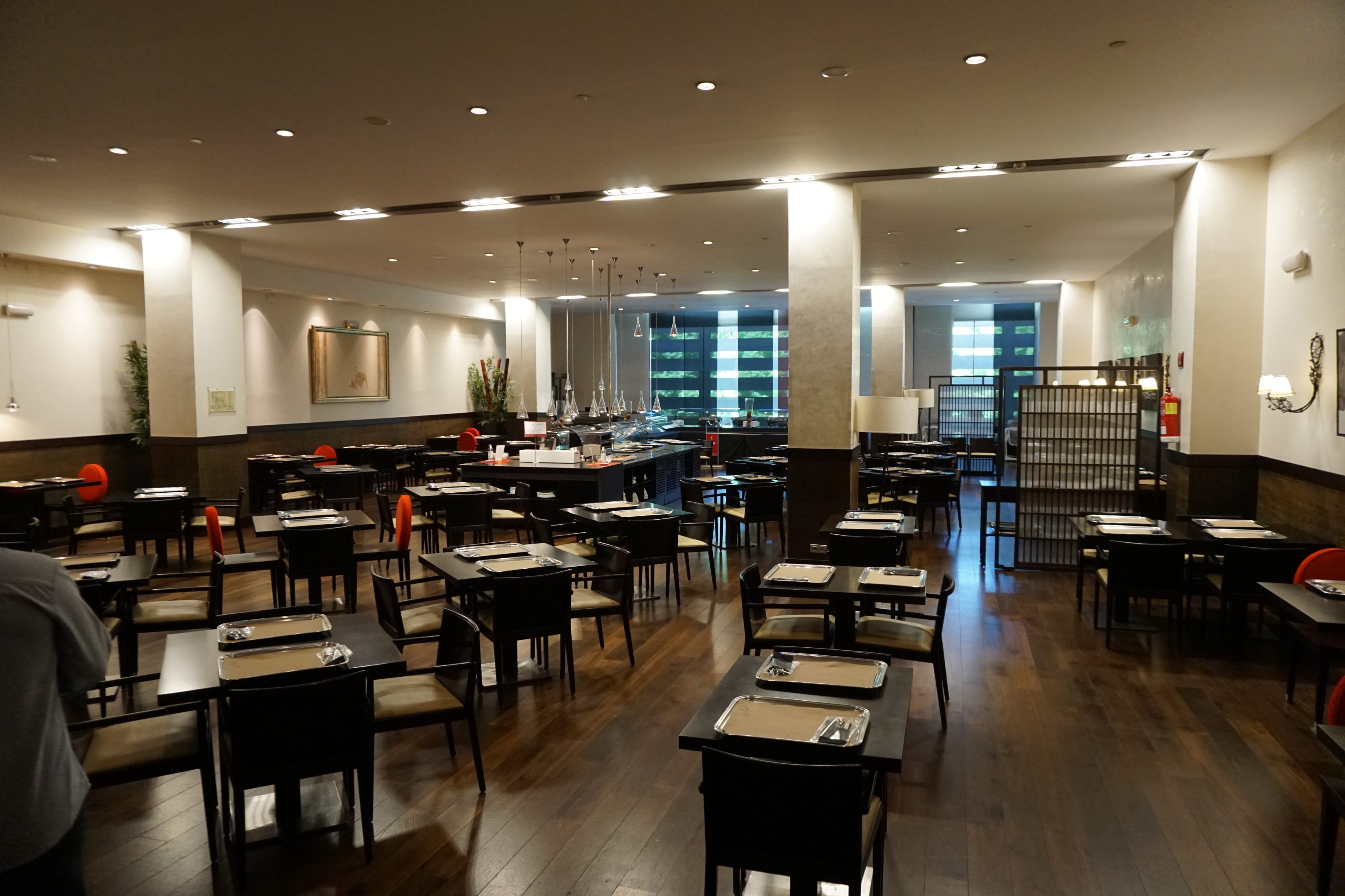 A modern restaurant interior with wooden flooring and neatly arranged dark wooden tables and chairs. Each table is set with silver trays. The lighting is dim and atmospheric, with several pendant lights hanging from the ceiling.